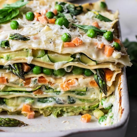 A close-up slice of Spring Veggie Alfredo Lasagna showcases tender zucchini, carrots, and peas nestled between rich layers of ricotta and melty mozzarella, garnished with fresh basil.